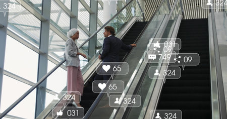 Talking business colleagues climbing escalator holding rail in glass atrium, with social icons. Corporate, teamwork, architecture, technology, networking, modern, collaborationの写真素材