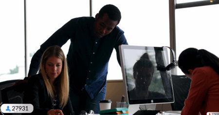 Reviewing documents, colleagues leaning over desk in modern office, with monitor and coffee cups. Collaboration, teamwork, workspace, professional, communication, productivity, modernの写真素材