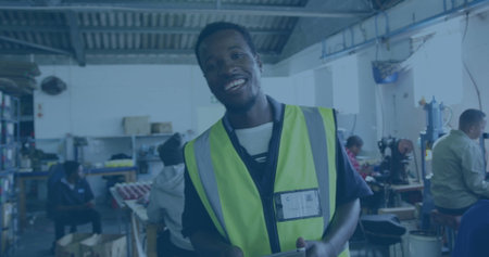Smiling African American worker wearing reflective vest holding tablet in workshop, with machinery....