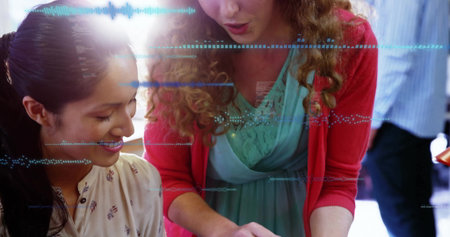 Leaning women collaborating over tablet at bright office desk, pointing at screen and documents....