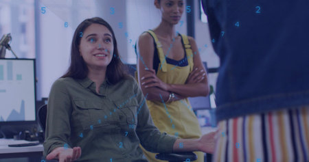 Woman in olive shirt pointing, explaining chart data in open plan office with monitors and overlays....