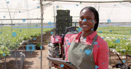 Holding tablet woman in red shirt apron inspecting lettuce in greenhouse, with tractor, copy space. Horticulture, sustainable farming, modern agriculture, urban farming, eco-friendlyの写真素材
