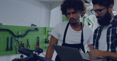 Two bike mechanics wearing aprons using laptop in workshop with handlebars, pegboard and shelving....