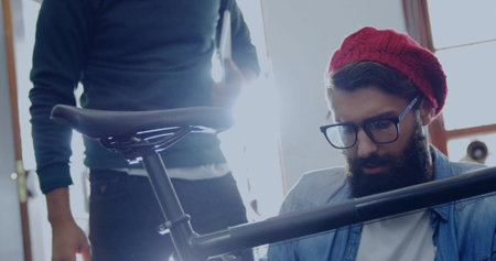Kneeling mechanic wearing red beanie adjusting seat post in workshop, with bicycle frame and saddle. Mechanic, collaboration, tools, craftsmanship, industrial, urban, teamworkの写真素材