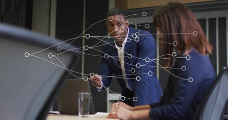 Leaning man in navy suit speaking across table in office, with laptop showing network overlay. Professional, collaboration, modern, communication, teamwork, corporate, networkingの写真素材