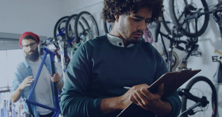 Curly-haired manager taking notes on clipboard inside bike workshop, with hanging frames and tools. Bike repair, maintenance, workshop, industrial, technical, team, craftsmanshipの写真素材