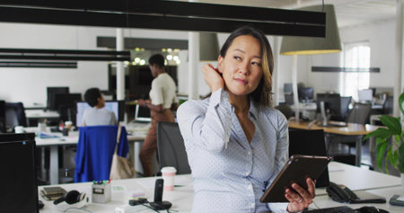 Mid adult Korean woman holding tablet and touching hair in open-plan office, with laptop coffee cup....
