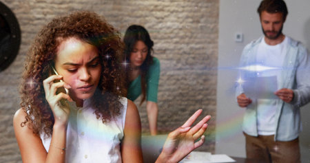 Holding smartphone, woman in blouse talking on phone at office meeting table with documents, sheets....