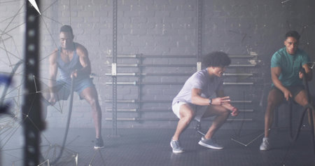 Instructor guiding clients wearing workout clothes waving battle ropes in gym, with barbell racks. Strength, endurance, fitness, training, athletic, muscular, powerの写真素材