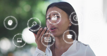 Speaking woman wearing white blouse holding voice device in park, displaying floating data icons....