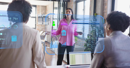 Presenting Asian woman in pink top gesturing toward data charts in meeting room with laptop, tablet....