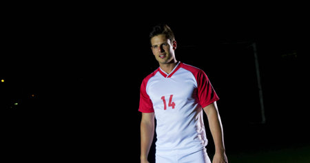 Soccer player standing in red-and-white jersey 14 on field at night, with goalpost, net, copy space. Athletic, teamwork, outdoor, competition, sport, training, performanceの写真素材