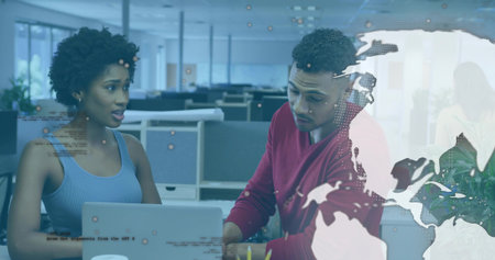 Colleagues wearing red sweater and blue top analyzing laptop in open office with digital overlays. Collaboration, technology, data visualization, digital workspace, professional, modern designの写真素材