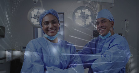 Smiling two surgeons wearing blue gowns posing in operating room, with overhead lights and monitors....