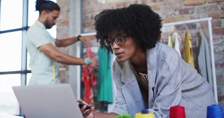 Female fashion designer leaning on desk holding stylus in loft studio, with laptop and tape measure. Creative, craftsmanship, industrial, workspace, inspiration, collaboration, eclecticの写真素材