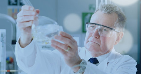 Scientist wearing lab coat and goggles examining glass flask at eye level in lab, with monitor. Research, innovation, chemistry, precision, science, experimentation, laboratoryの写真素材