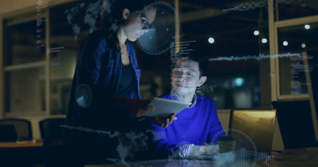 Colleagues wearing denim jacket reviewing on laptop and tablet in office at night, with overlays. Technology, collaboration, innovation, data visualization, teamwork, modern, professionalの写真素材