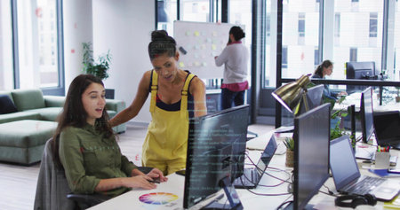 Collaborating female designers pointing at monitors in open-plan office, with printed color wheel. Collaboration, creativity, modern, workspace, innovation, teamwork, productivityの写真素材