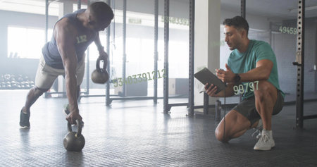 Performing renegade row man holding two kettlebells at gym with trainer tablet, copy space....
