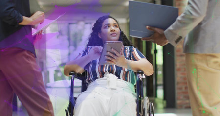 Gesturing wheelchair user showing tablet to colleagues with clipboard and laptop in office corridor....