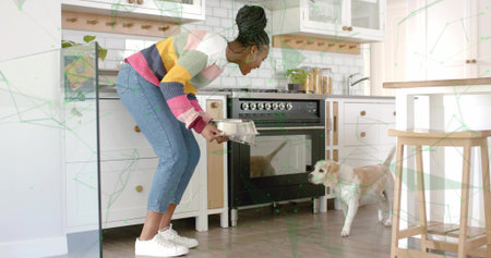 Bending woman wearing striped top holding stainless steel dog bowl in kitchen with Labrador puppy. Interior, pet, modern, cozy, lifestyle, home decor, companionshipの写真素材