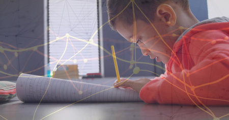 Boy in red sweatshirt writing in notebook at desk by blinds, with yellow pencil, network overlay. Student, education, creativity, concentration, learning, technology, innovativeの写真素材