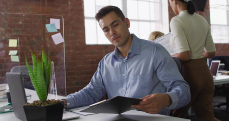 Reviewing black folder, man wearing blue shirt sitting at office desk, with acrylic divider....