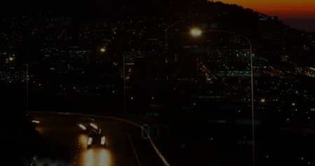 Driving two cars down hillside road at dusk, with headlights, streetlamp, guardrail, copy space....