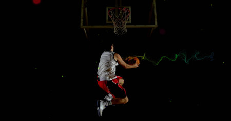 Leaping male basketball player dunking basketball into hoop on dark court, wearing white jersey....