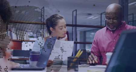 Collaborating colleagues in blue blouse and red shirt using laptop and reports in open-plan office. Modern, workplace, teamwork, professional, productivity, communication, corporateの写真素材