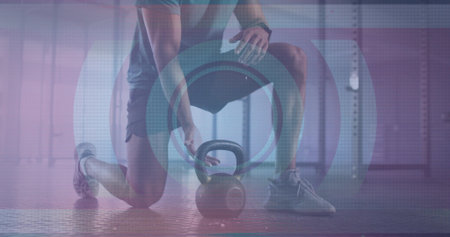 Kneeling athlete wearing athletic shirt gripping kettlebell on rubber gym floor, weight rack behind. Fitness, strength, training, gym, endurance, durability, athleticismの写真素材