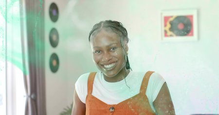 Smiling woman wearing rust pinafore dress posing in studio with curtains, plant, records, artwork....