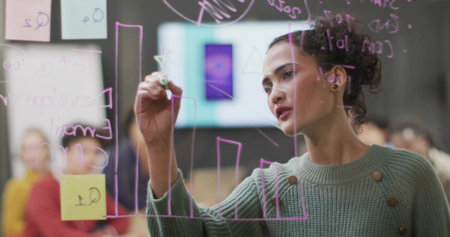 Presenting Hispanic woman writing purple charts on glass board in boardroom, with sticky notes. Business, strategy, innovation, collaboration, modern, teamwork, leadershipの写真素材