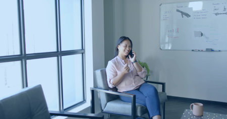 Talking Asian woman holding smartphone gesturing in armchair at office lounge with whiteboard....