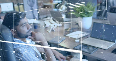 Leaning South Asian man processing network data at modern office desk, with laptop, monitors, plant. Technology, innovation, workspace, digital, analysis, software, productivityの写真素材