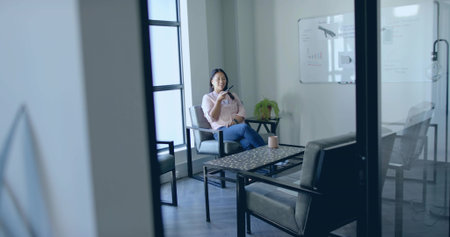 Talking businesswoman speaking into smartphone in meeting lounge, with ceramic mug, copy space. Corporate, workspace, modern, casual, communication, interior, professionalの写真素材