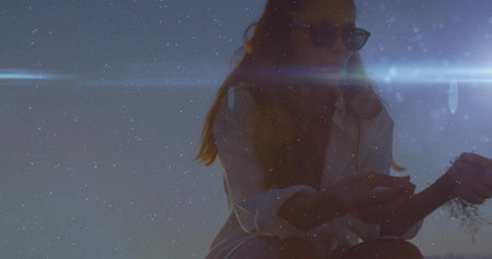 Examining dried plant, woman in white shirt, sunglasses under lens-flare streak on dune, copy space....