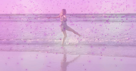 Balancing woman in short floral dress lifting leg in shallow beach water, with pink dot overlay. Summertime, serenity, vibrancy, nature, relaxation, elegance, leisureの写真素材