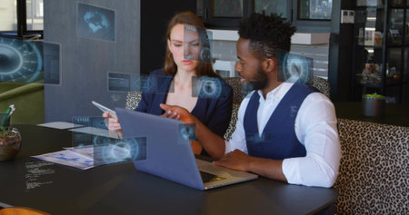 Reviewing data two business professionals pointing at laptop and tablet in office, with holograms....