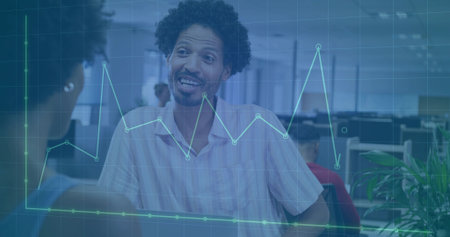 Curly-haired man talking to female colleague in open-plan office, with monitors and chart overlay....