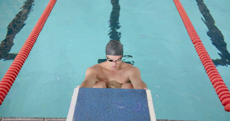 Crouching swimmer bracing on blue starting block at pool edge, with black swim cap and goggles. Athlete, competition, aquatic, aquatic sports, training, fitnessの写真素材