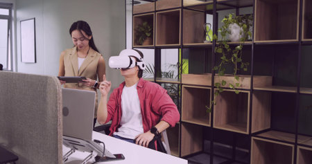 Man in red overshirt testing virtual reality headset at office desk, with tablet laptop shelf....
