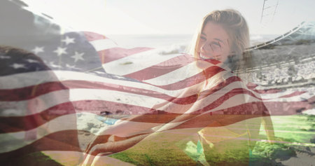 Smiling woman in yellow top sitting on grass facing man, with American flag overlay and bleachers. Patriotism, leisure, coastal, outdoor, happiness, summer, relaxationの写真素材