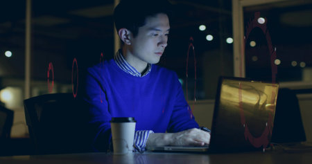 Working Chinese man in blue sweater typing on laptop late night in office, with coffee cup....