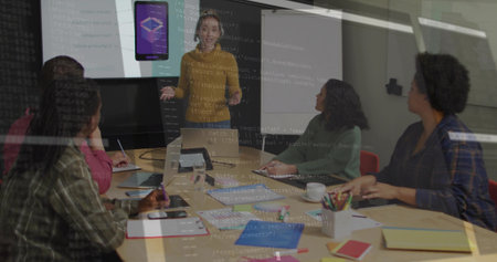 Presenting woman in mustard top pointing at app display in meeting room, with laptops, notebooks....