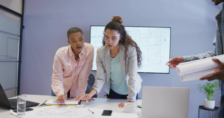Discussing professionals leaning over blueprints in conference room, with rolled plans and screen....