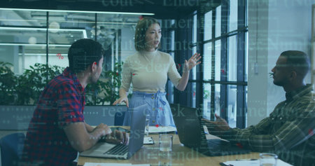 Female colleague wearing business attire gesturing in meeting room with three laptops and notebooks....