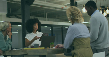 Gesturing professional woman holding tablet around high table in open-plan office, with laptop pens....