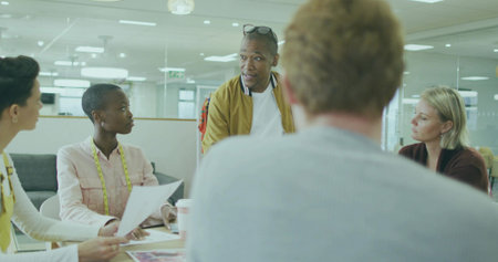 Addressing man in brown jacket speaking at office table with printed documents and measuring tape....