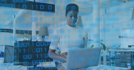 Working woman wearing white t-shirt typing on laptop in open-plan office, with monitor and plant....